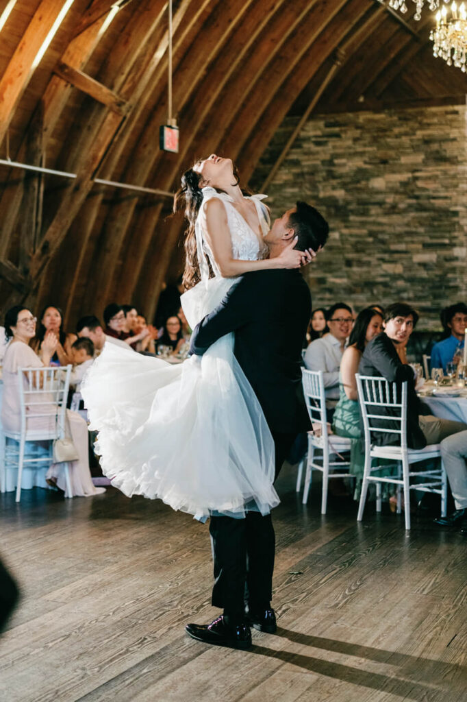 The groom lifts his bride into the air during their first dance, her tulle gown floating gracefully as she leans back in laughter, surrounded by guests at Sweeney Barn.