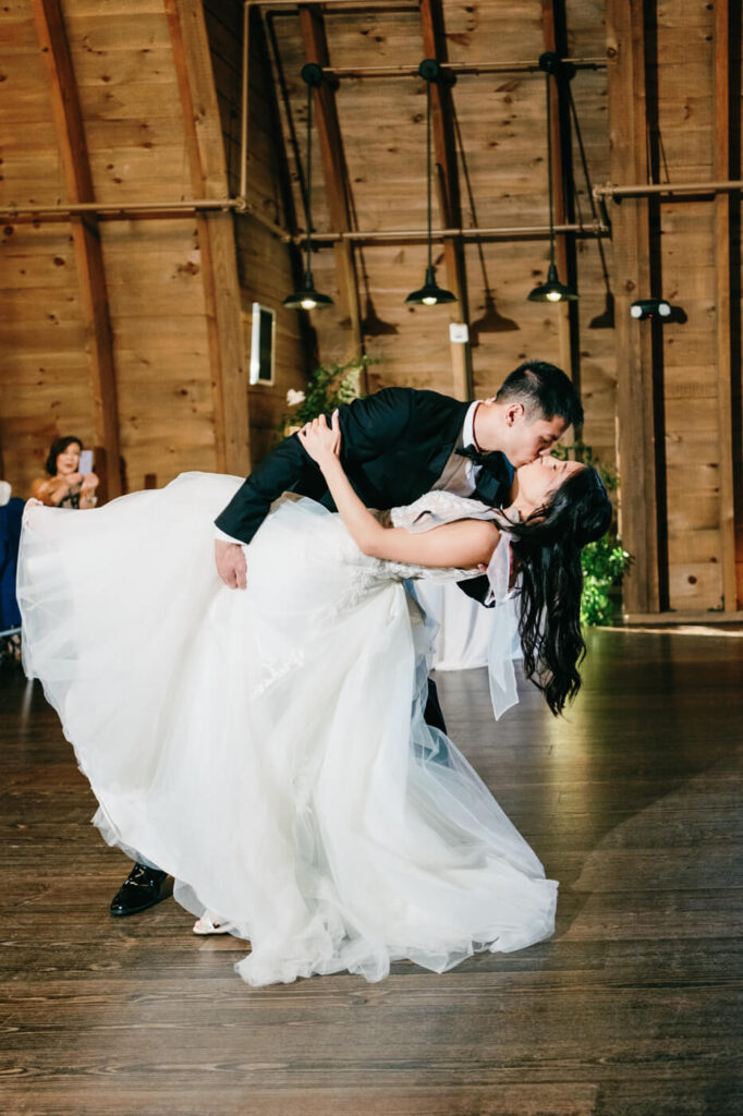 A romantic moment as the groom dips his bride for a kiss during their first dance, her wedding dress flowing elegantly across the dance floor beneath the barn’s warm lighting.