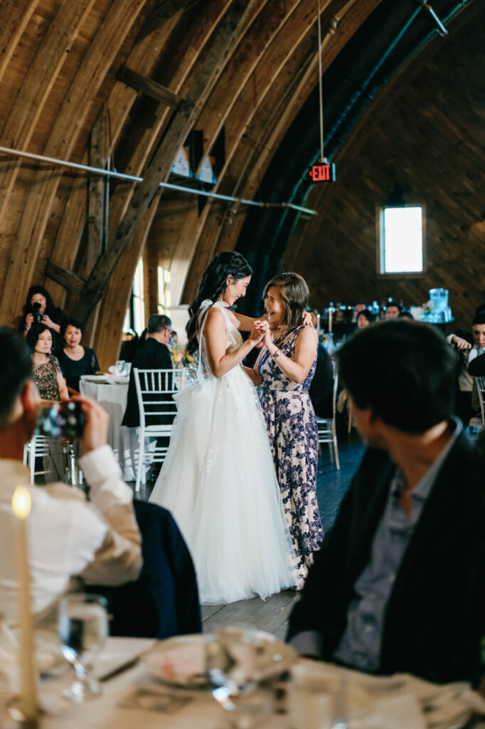 The bride shares a heartfelt dance with her mother, both smiling tenderly as they hold hands on the reception floor, while guests watch in the background.