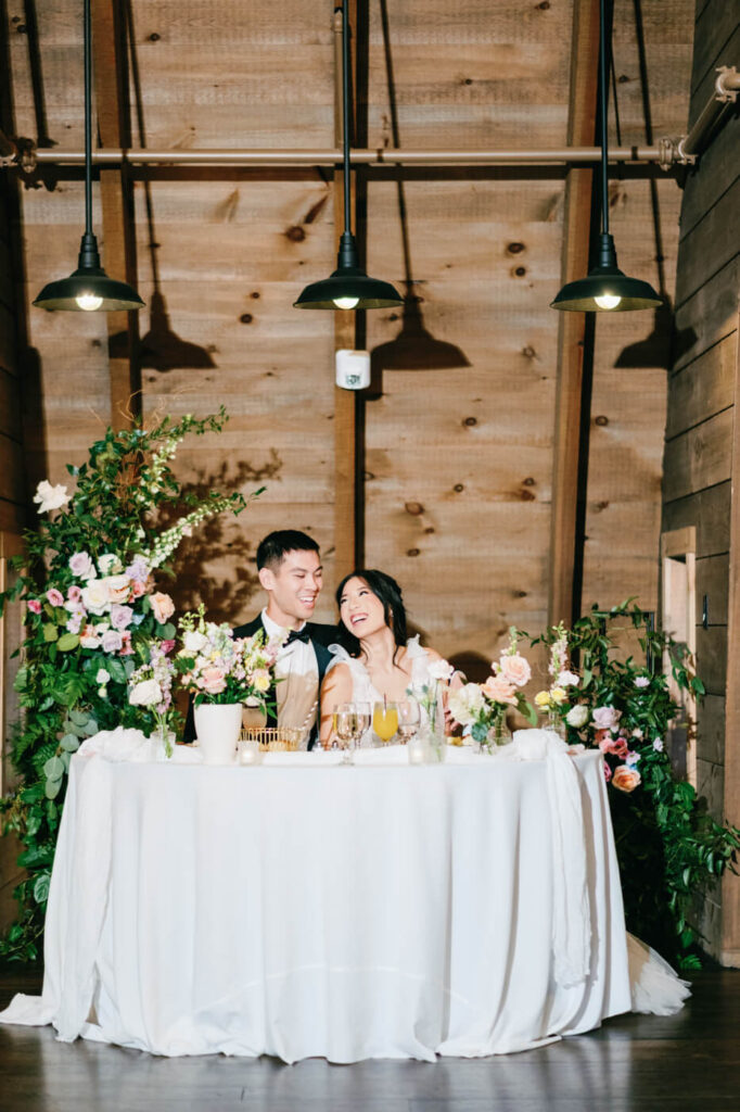 The bride and groom sit together at their sweetheart table, surrounded by lush floral arrangements and glowing candlelight, sharing a joyful moment as they look at each other with laughter.