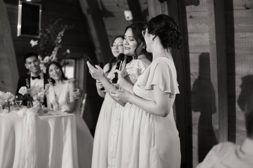 A bridesmaid & sister gives a heartfelt toast to the newlyweds, reading from her phone as she holds back tears, while the bride and groom smile warmly in the background.