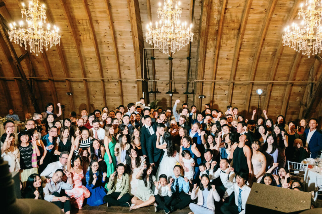 A celebratory group photo of all the wedding guests surrounding the bride and groom, cheering and raising their hands in the beautifully lit barn reception space.