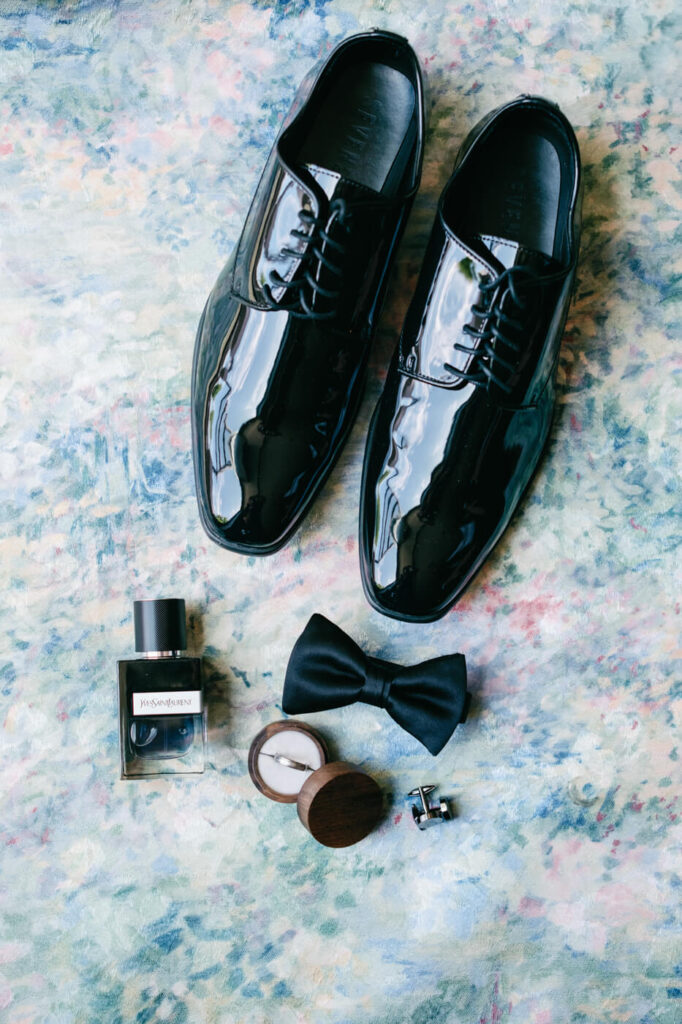 Groom’s Wedding Accessories – Black patent leather shoes, bowtie, YSL cologne, and wedding band arranged on a pastel floral fabric, highlighting the groom’s wedding day details at Sweeney Barn.