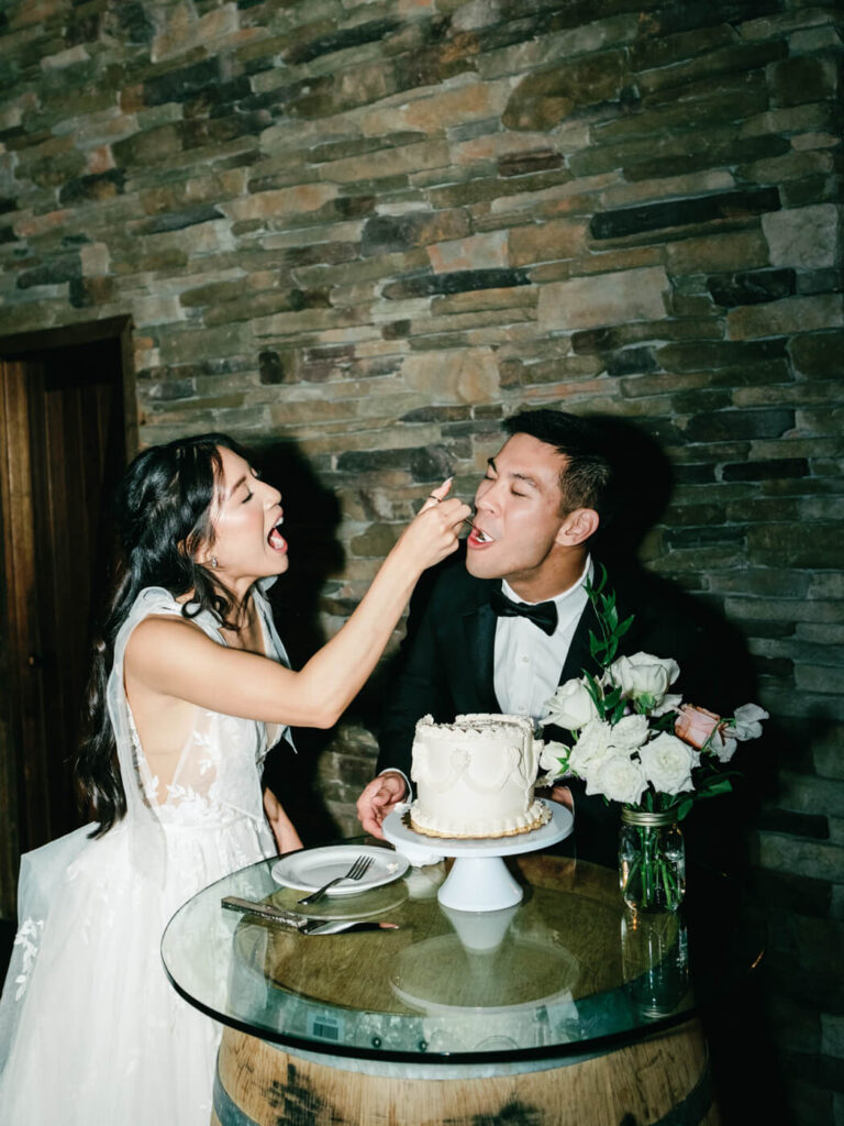 The bride playfully feeds a bite of cake to her groom as they laugh together during the cake-cutting moment, standing beside a rustic barrel table adorned with florals and candlelight.