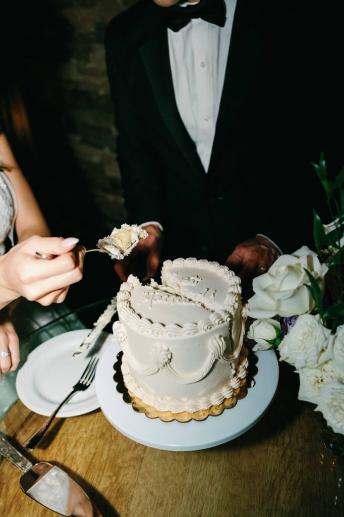 A bride and groom cut their wedding cake together, with the bride holding a forkful of cake. The elegant white cake sits on a wooden table, surrounded by white roses.