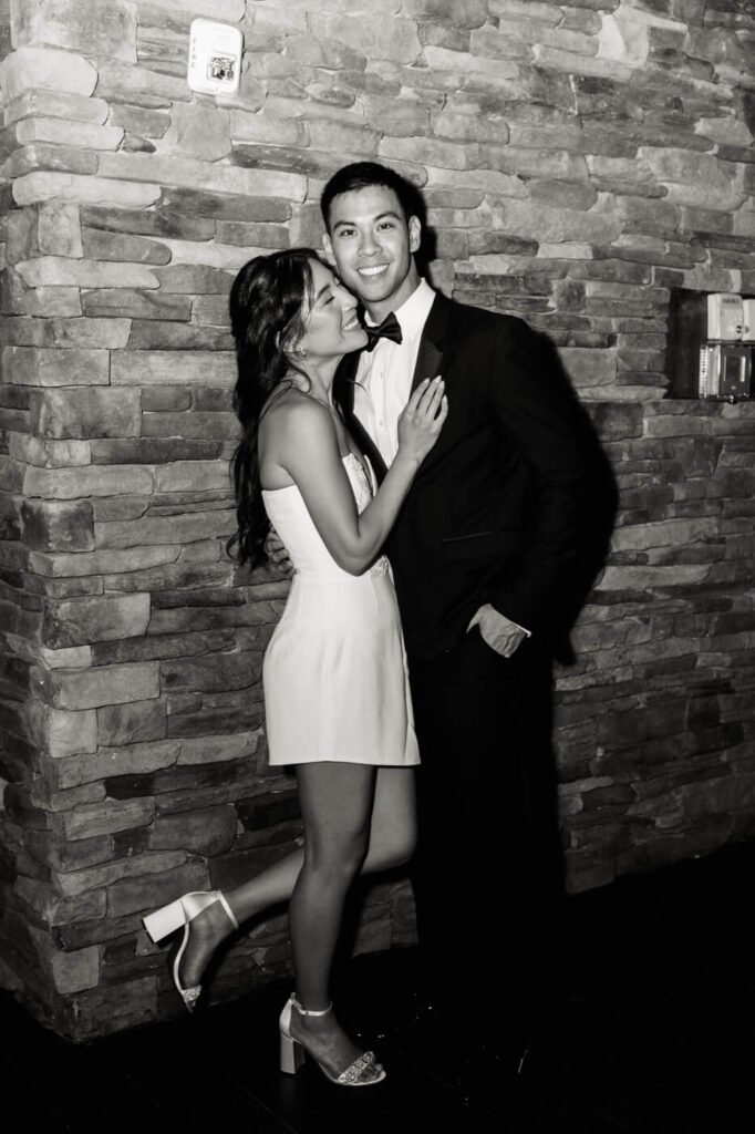 A black-and-white photo of a bride in a short white dress and a groom in a tuxedo embracing by a stone wall. The bride leans in with a joyful smile while the groom looks at the camera.