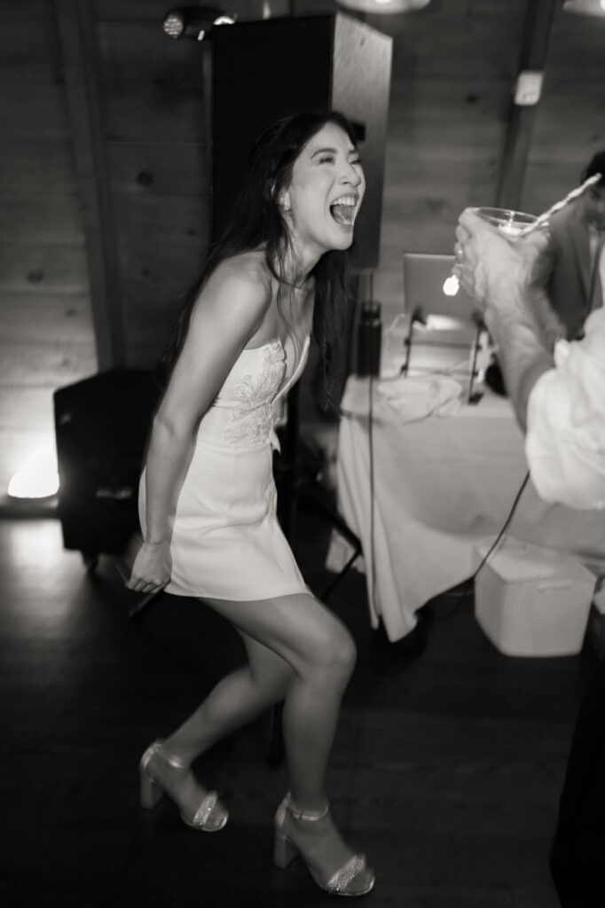 A black-and-white candid of a bride in a white mini dress laughing wholeheartedly on the dance floor, holding a drink as she enjoys the celebration.