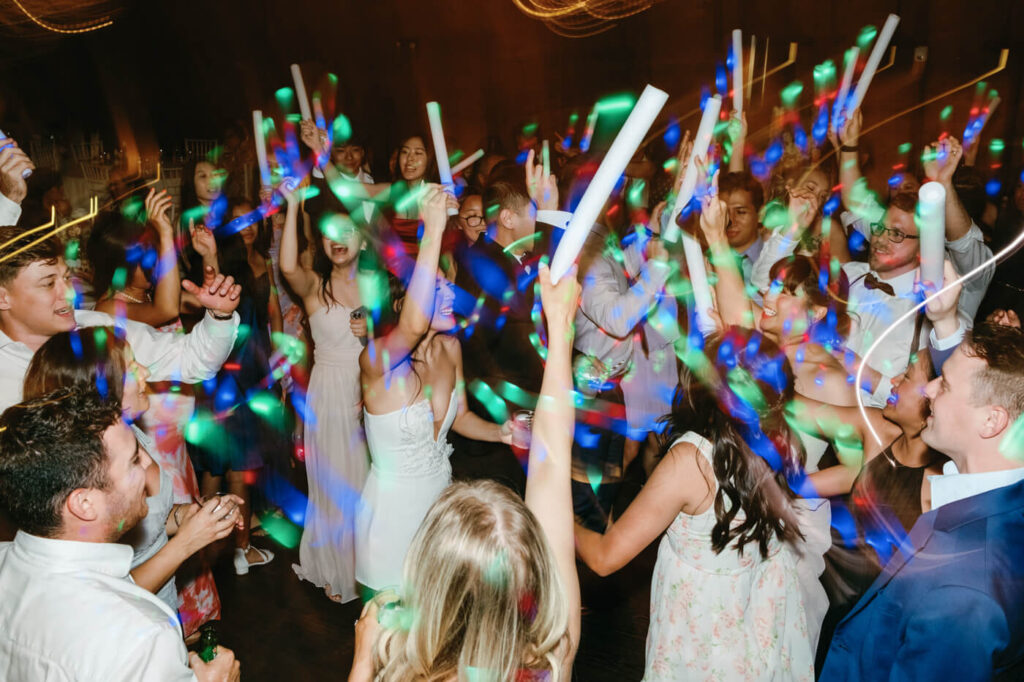 A lively wedding reception scene with guests dancing under colorful lights, holding glow sticks in the air, creating an atmosphere of high-energy celebration.