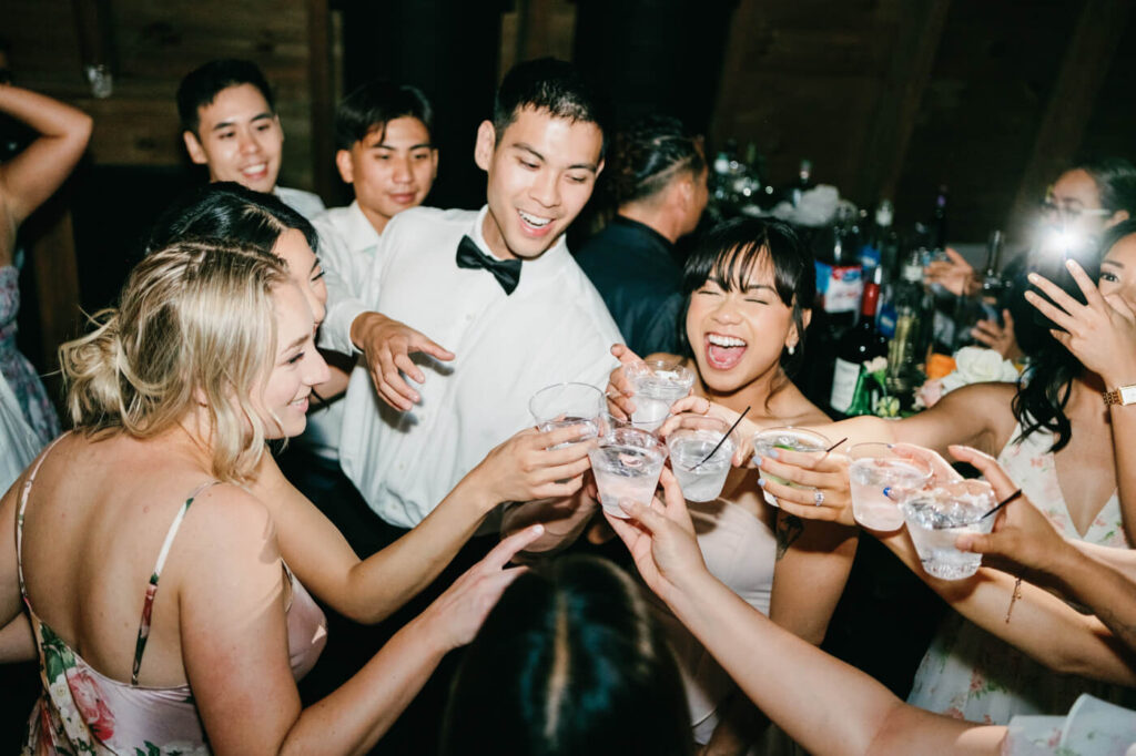 The groom, in a white dress shirt and bowtie, toasts with wedding guests, all smiling and raising their glasses in celebration.