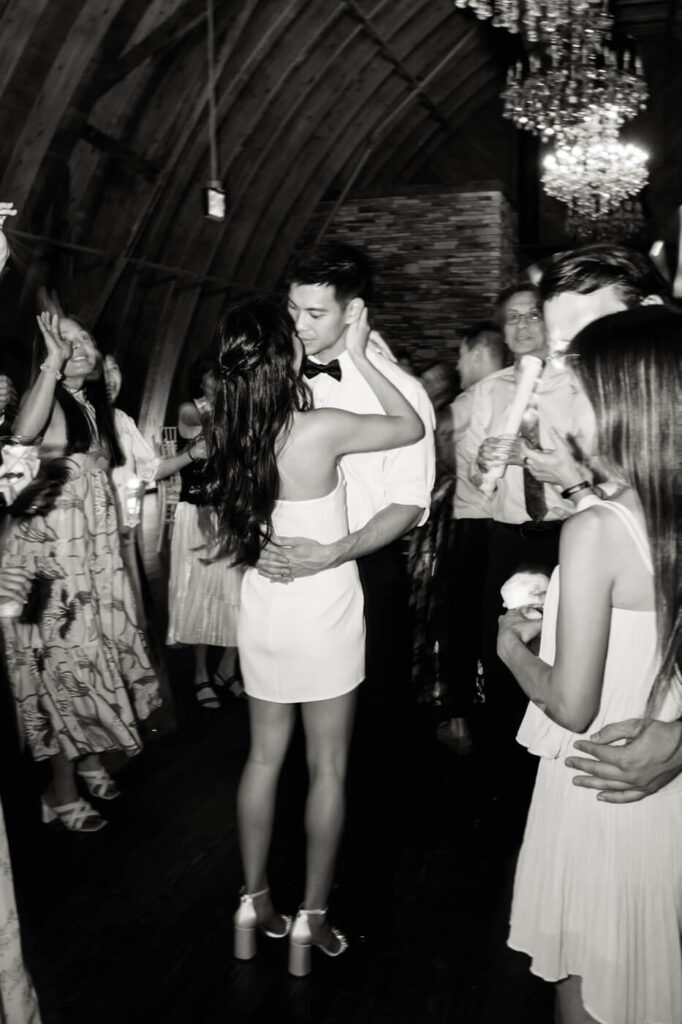 A black-and-white image of the couple sharing a romantic moment on the dance floor. The bride, in a short white dress, embraces the groom while guests celebrate around them.