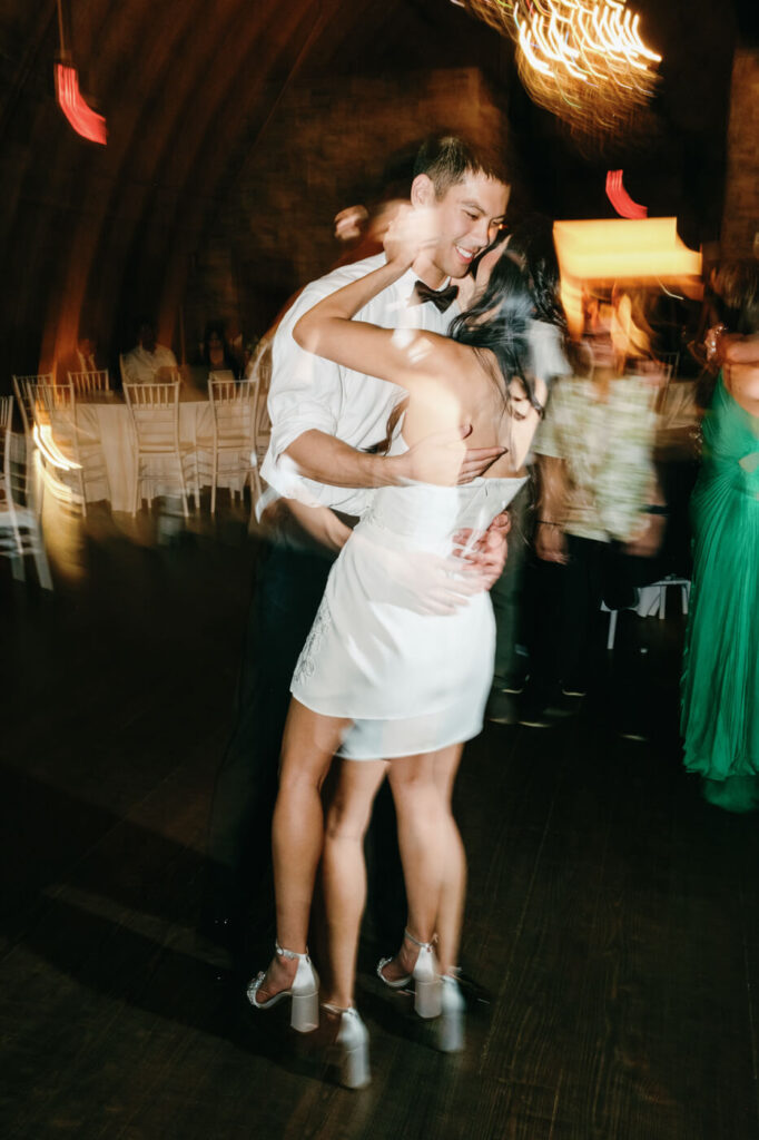 The bride and groom dance closely, smiling at each other under warm golden lighting, with motion blur capturing the energy of the moment.