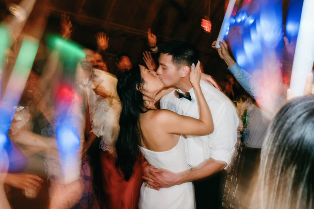 A dramatic, motion-filled image of the couple sharing a kiss in the middle of a lively dance floor, surrounded by guests celebrating with glow sticks and colorful lighting.