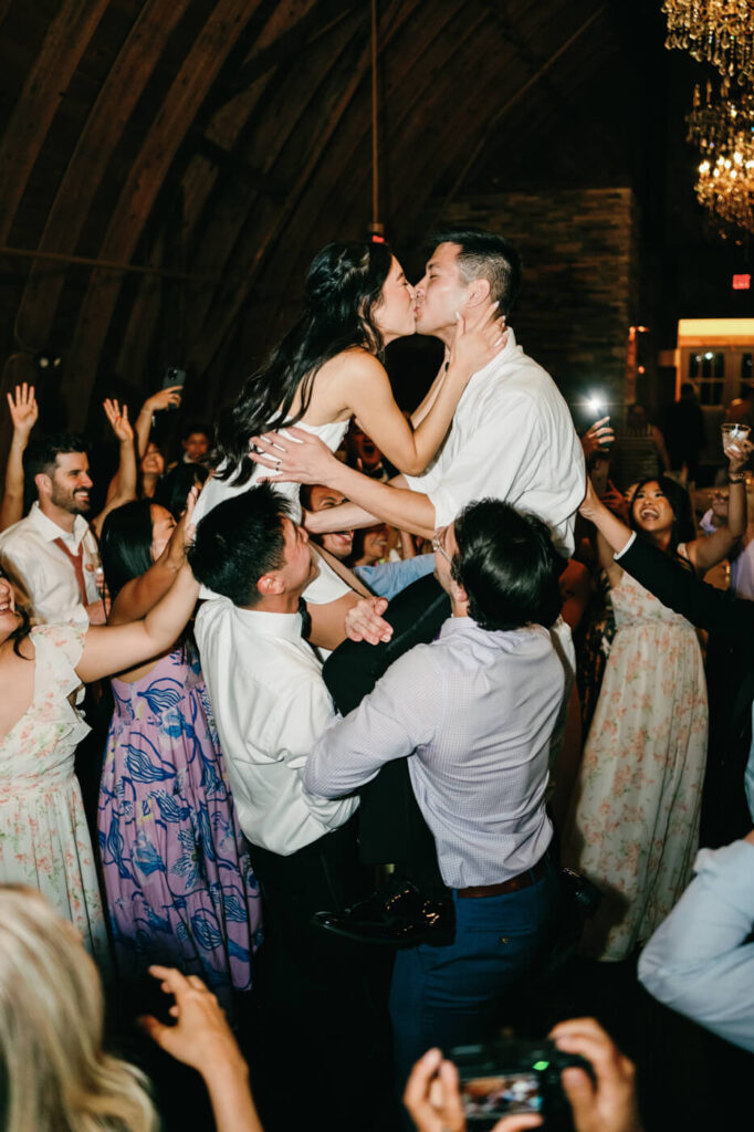 The groom is lifted into the air by friends while kissing the bride, who is also being held up, creating an electrifying and joyful wedding party moment.