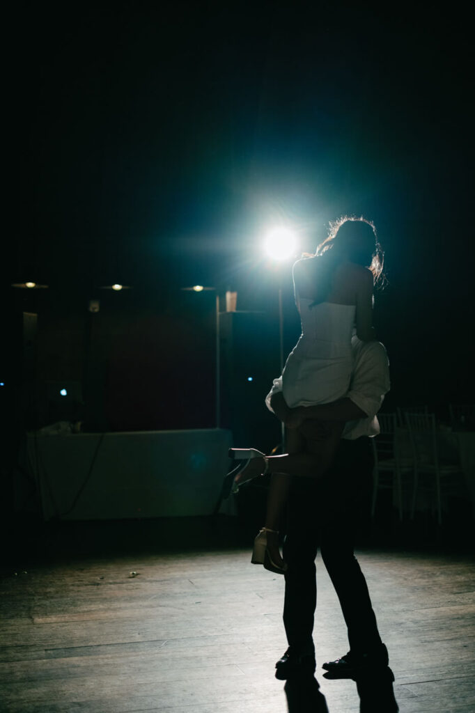 A moody, artistic silhouette of the groom lifting the bride in the air on the dance floor, with a bright spotlight behind them, capturing a cinematic and romantic moment.