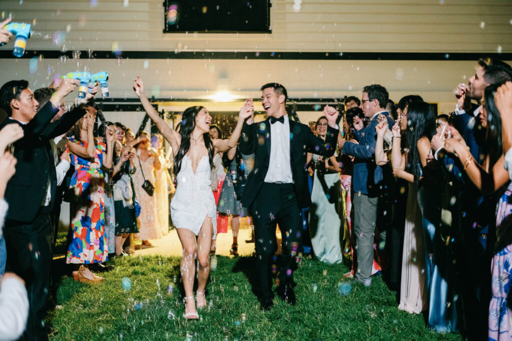 Lively image of the bride & groom exiting the Sweeney Barn amidst their guests surrounded by bubbles during their bubble grand exit for a celebratory moment.