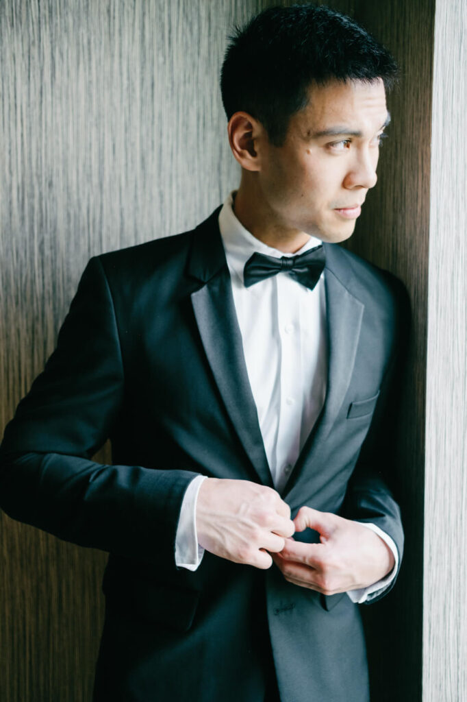 Groom in Tuxedo by Window – Groom gazes out the window while buttoning his tuxedo jacket, anticipating his wedding day at Sweeney Barn in Virginia.