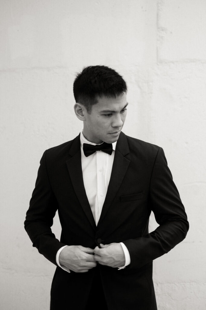 Black and White Portrait of Groom – Classic black and white image of the groom adjusting his suit against a minimal white wall, evoking timeless elegance at Sweeney Barn.