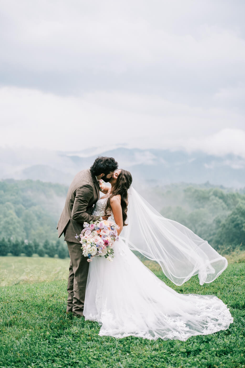 Bride and groom sharing a kiss in the rain with mountains in the background at an Asheville mountain wedding by The Reeses.