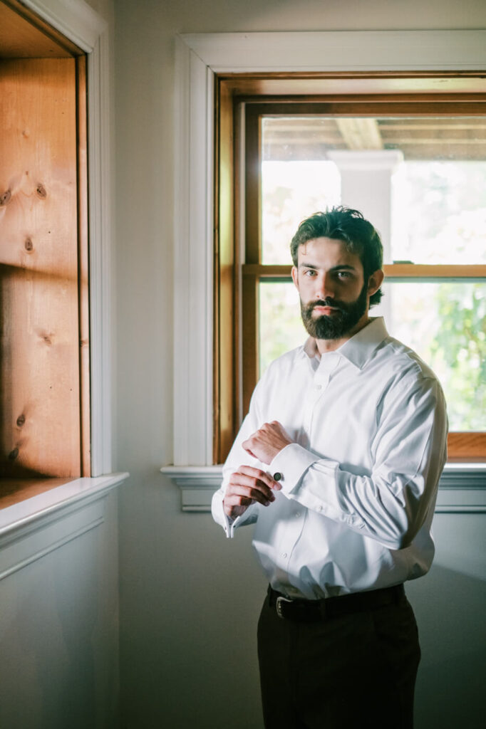 Groom adjusting cufflinks by a window, part of an Asheville mountain wedding captured by The Reeses.