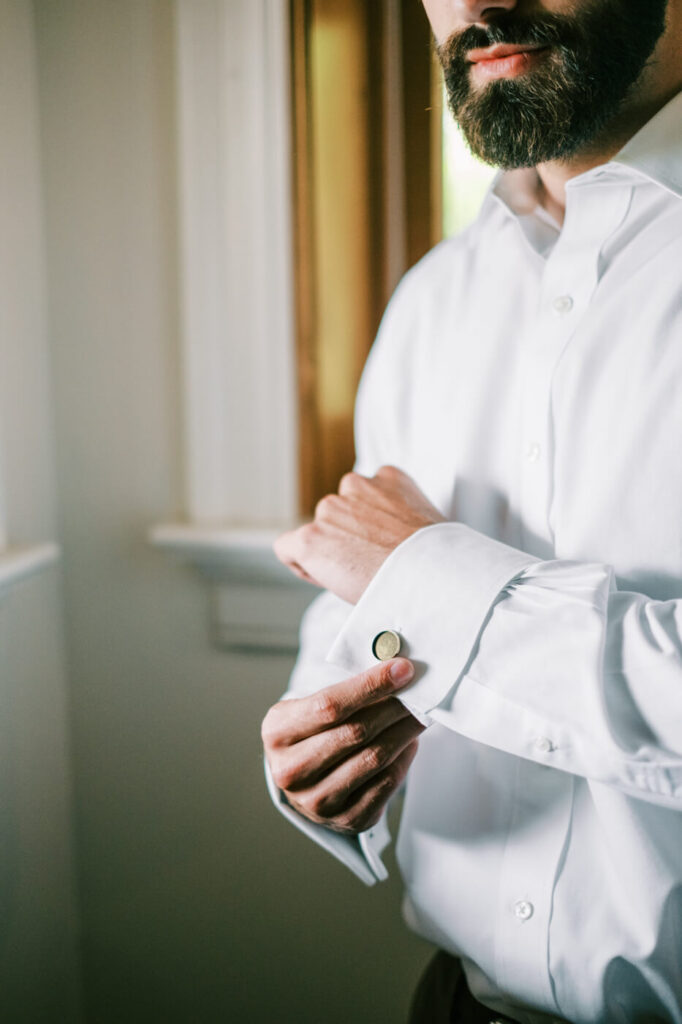 Groom adjusting cufflinks by a window, preparing for an Asheville mountain wedding captured by The Reeses.