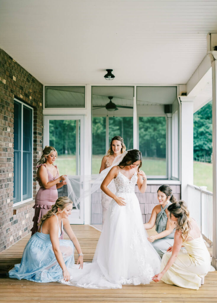 Bridesmaids in pastel dresses adjusting bride’s veil and dress on a porch, part of an Asheville mountain wedding by The Reeses.