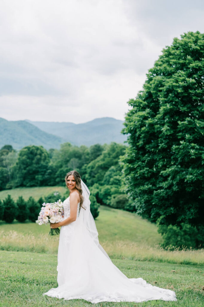 Bride with pastel bouquet in a field with mountains, part of an Asheville mountain wedding photographed by The Reeses