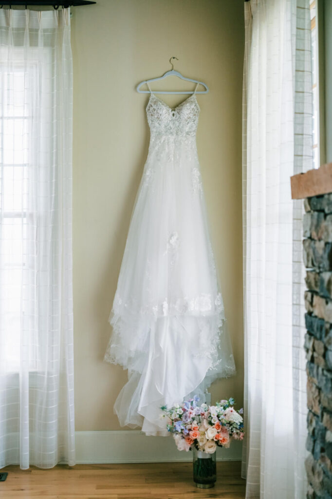 Elegant white wedding dress with lace details hanging in a bright room for an Asheville mountain wedding by The Reeses.