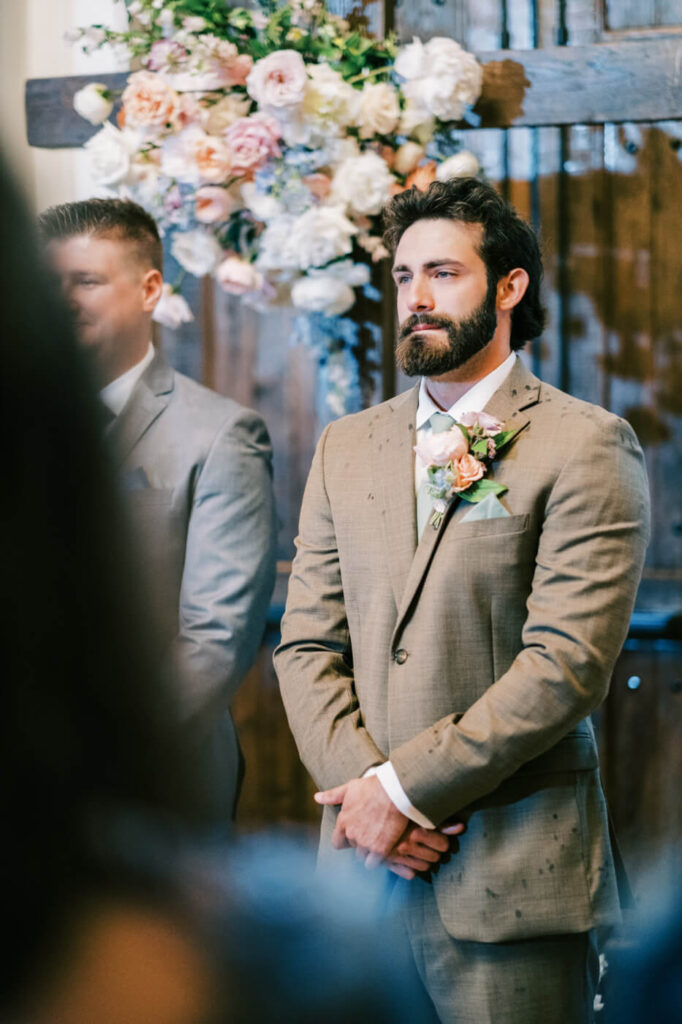 Groom waiting for bride during indoor ceremony with floral backdrop, part of an Asheville mountain wedding by The Reeses.