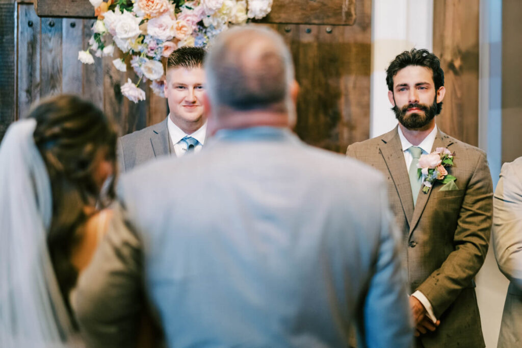 Groom watching bride walk down the aisle standing under floral cross, part of an Asheville mountain wedding captured by The Reeses.