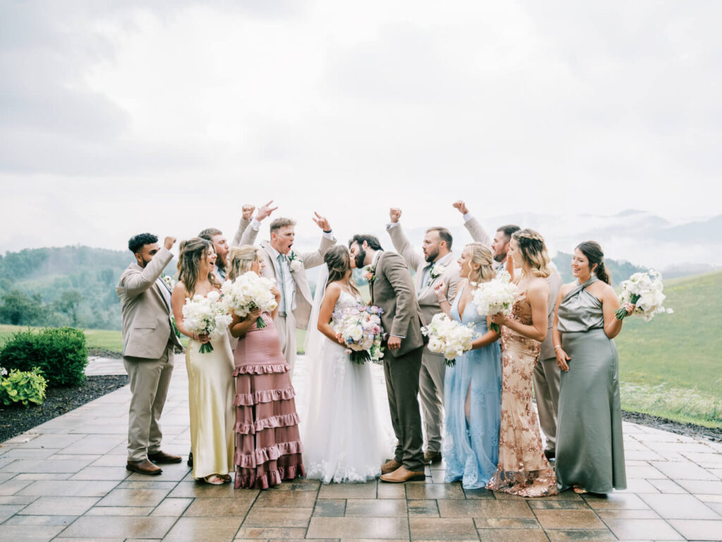 Wedding party cheering with bouquets and mountains as bride & groom kiss, part of an Asheville mountain wedding by The Reeses.