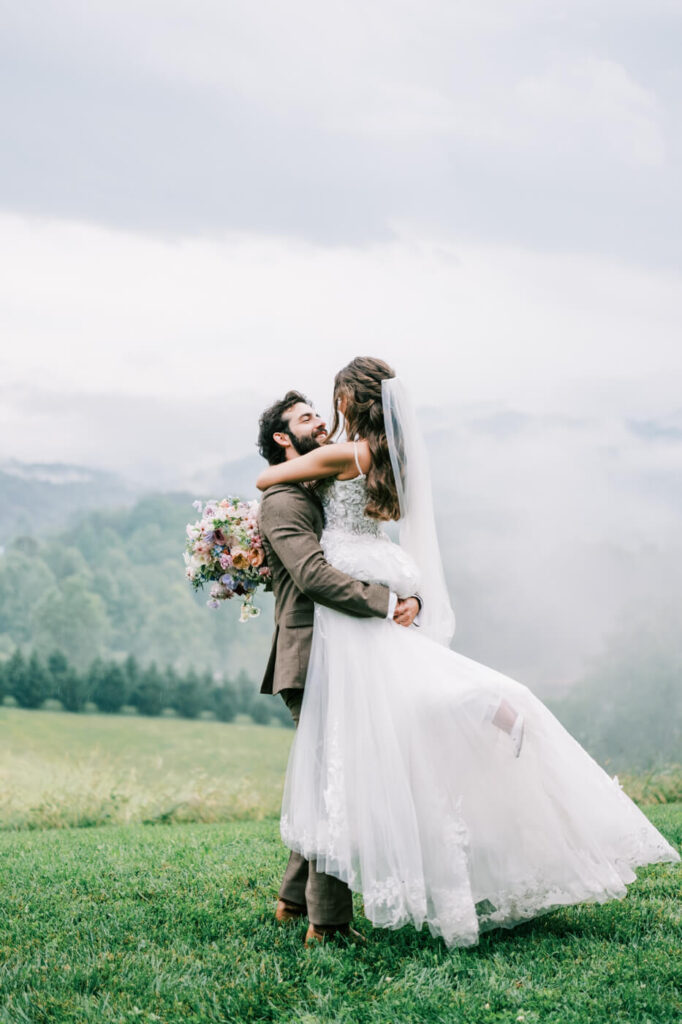 Bride and Groom Dance in the Rain at Asheville Wedding – The groom twirls the bride in the rain, her flowing gown billowing, with dreamy blue-gray mountain mist in the background.