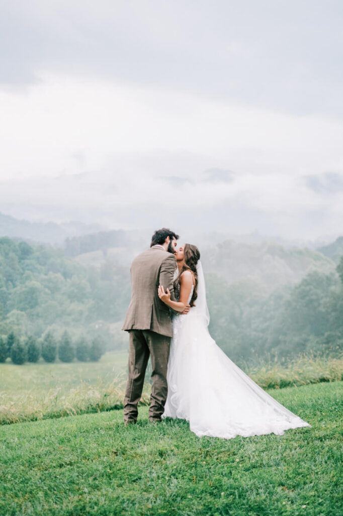 Bride and Groom Kiss in Asheville Mountains – A candid moment as the newlyweds kiss with rolling foggy hills creating a cinematic and romantic atmosphere.