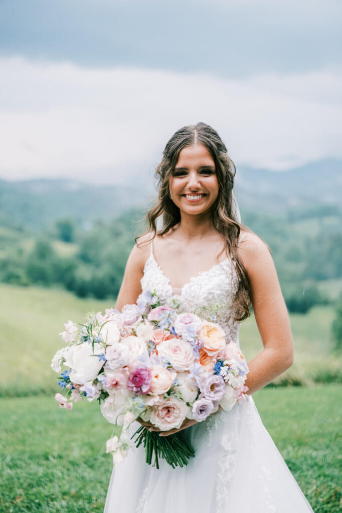 Bride Holding Lush Pastel Bouquet at Asheville Wedding – A joyful bride smiles while holding a cascading bouquet of pastel roses, peonies, and wildflowers, with rolling North Carolina mountains behind her.