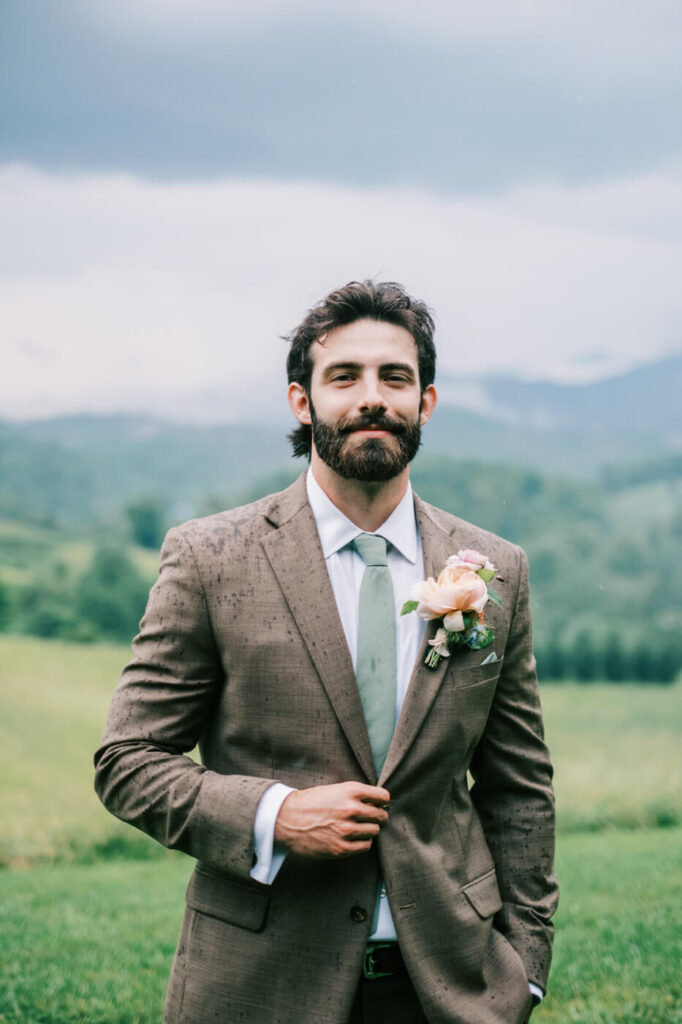 Groom Portrait in Rain at Asheville Wedding – The groom, dressed in a tailored brown suit and sage green tie, stands confidently with raindrops on his jacket, embodying rustic elegance against the mountain backdrop.