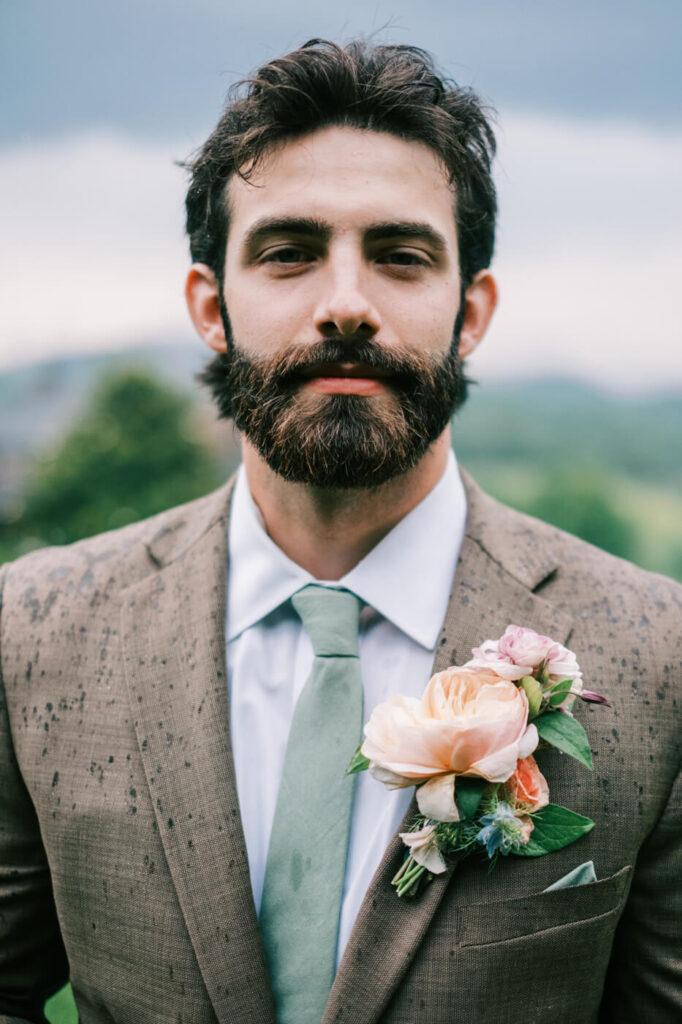 Close-up of Groom’s Wedding Boutonniere and Suit – A detailed shot of the groom’s floral boutonniere featuring a blush rose, set against his textured brown suit, with misty Asheville mountains behind.