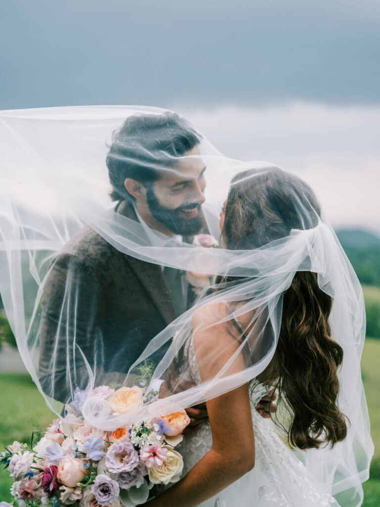 Bride and Groom Under Wedding Veil in Asheville Mountains – A romantic, intimate moment as the bride and groom gaze at each other under her flowing veil, framed by the moody North Carolina mountain skies.