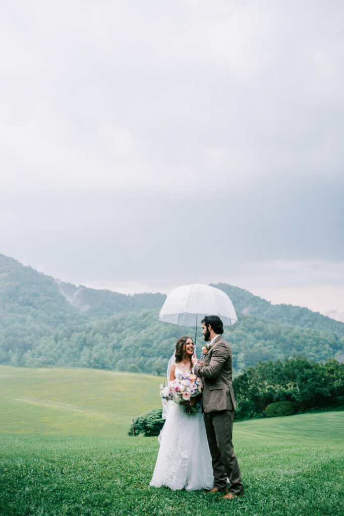 Bride and Groom’s Rainy Mountain Wedding Moment with Umbrella – The couple stands in the lush Asheville hills, embracing under a clear umbrella, proving that a little rain only adds to the romance.