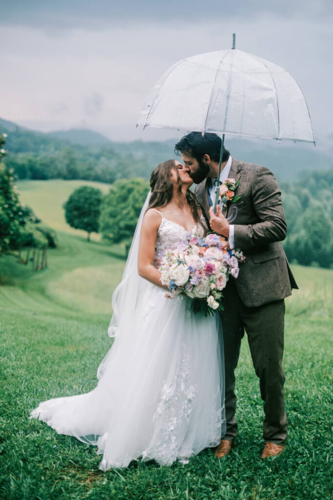 Bride and Groom’s Rainy Mountain Wedding Moment with Umbrella – The couple kisses in the lush Asheville hills, under a clear umbrella, proving that a little rain only adds to the romance.