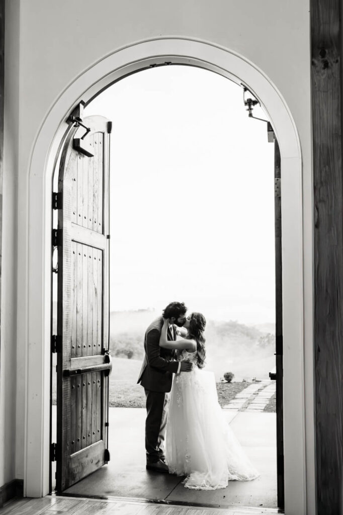 A bride and groom share a kiss framed by an elegant arched doorway, with misty mountain views in the background. Captured at a luxury Asheville wedding venue.