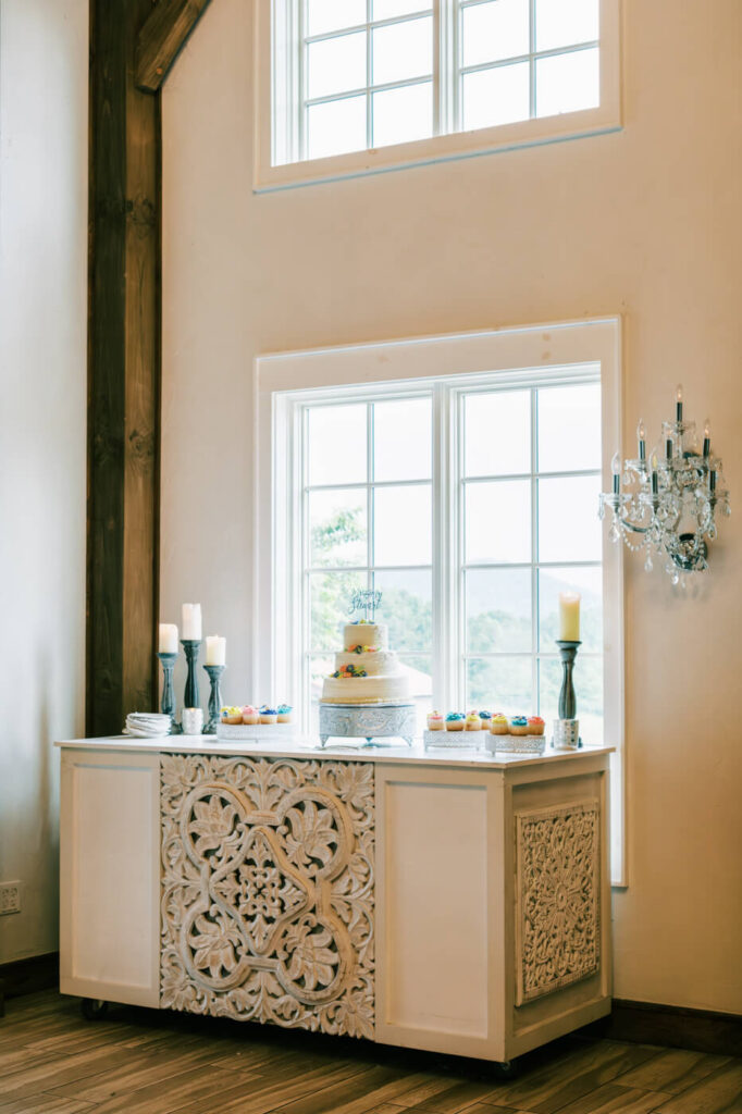 Asheville Wedding Cake Display with Vintage Elegance – A wedding cake and dessert table adorned with ornate white cabinetry, romantic candlelight, and pastel cupcakes, creating a refined reception scene.