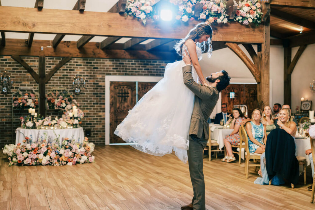 Bride and Groom’s First Dance in Rustic Asheville Venue – The groom lifts the bride joyfully during their first dance, surrounded by romantic florals and candlelit tables inside a rustic-luxury wedding venue.