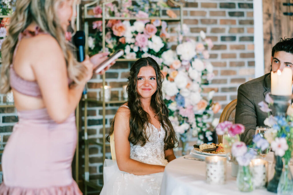 Bride’s Heartfelt Reaction to Wedding Toasts – The bride smiles warmly while listening to touching wedding toasts, seated at her elegantly decorated reception table in Asheville, NC.