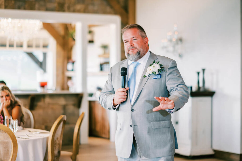 Father of the Bride Wedding Speech in Asheville Venue – The father of the bride shares a moving speech during the wedding reception, standing in front of a softly lit and elegant dining space.