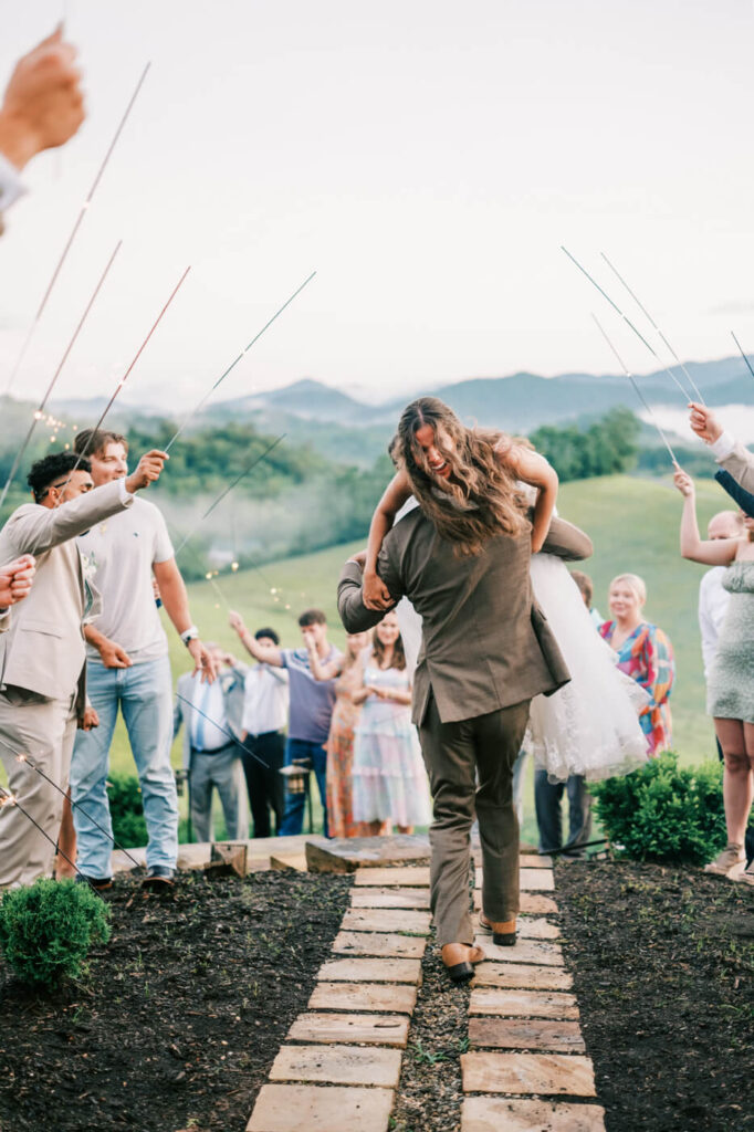 Bride carried by groom during sparkler exit in Asheville wedding – The groom playfully carries his bride as they make a grand exit through a pathway of sparklers, with the scenic Asheville mountains in the background.