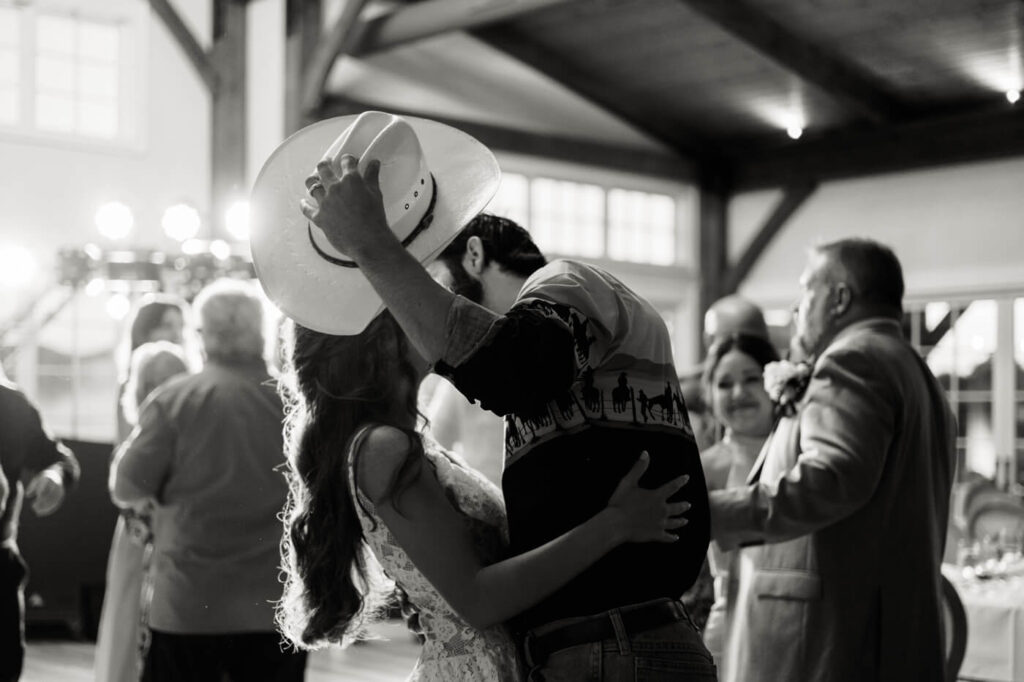 Groom kisses bride while tipping cowboy hat during wedding dance – The groom pulls his bride close for a passionate kiss, his cowboy hat adding a stylish Western touch to this intimate wedding dance moment.