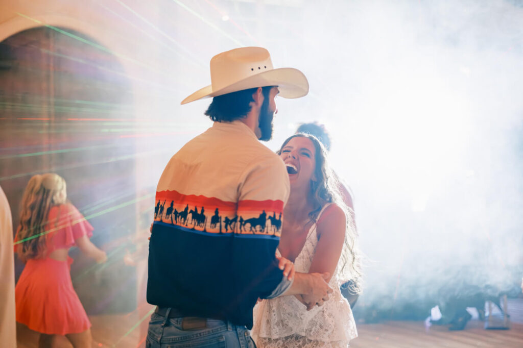 Bride and groom sharing a joyful dance – The bride laughs joyfully while holding hands with her groom under glowing lights, with soft smoke and colorful beams creating a magical wedding reception moment.