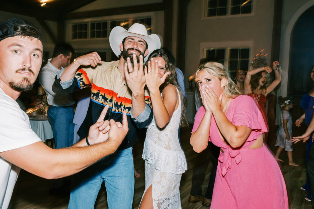 Wedding couple celebrating with guests – The groom proudly flashes his wedding band, standing beside his bride as their friends react with excitement during their fun-filled Asheville wedding reception.