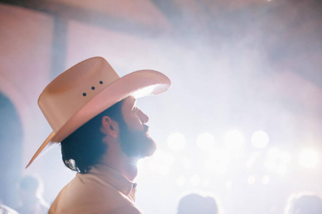 Groom in cowboy hat under wedding reception lights – A groom in a white cowboy hat gazes toward the dance floor, illuminated by dreamy light beams during his mountain wedding celebration.