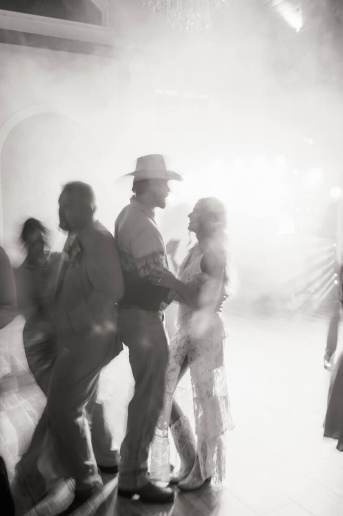 Western wedding dance floor moment – A groom in a cowboy hat and his bride are silhouetted in soft wedding reception lighting, dancing together amid a dreamy haze.