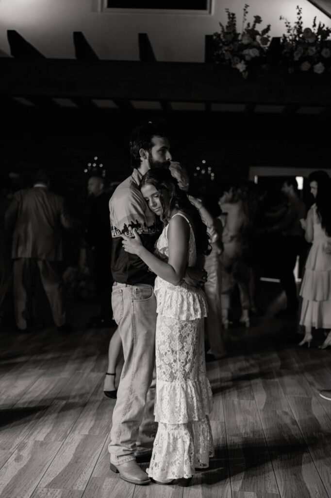 Bride and groom sharing an intimate slow dance – A black-and-white photo of the bride resting her head on her groom’s chest as they embrace on the dance floor at their rustic barn wedding.